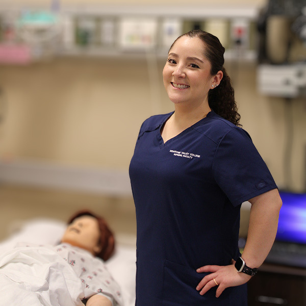 Carolina Martinez Calderon wears dark blue scrubs and stands next to a manikin in the nursing lab.