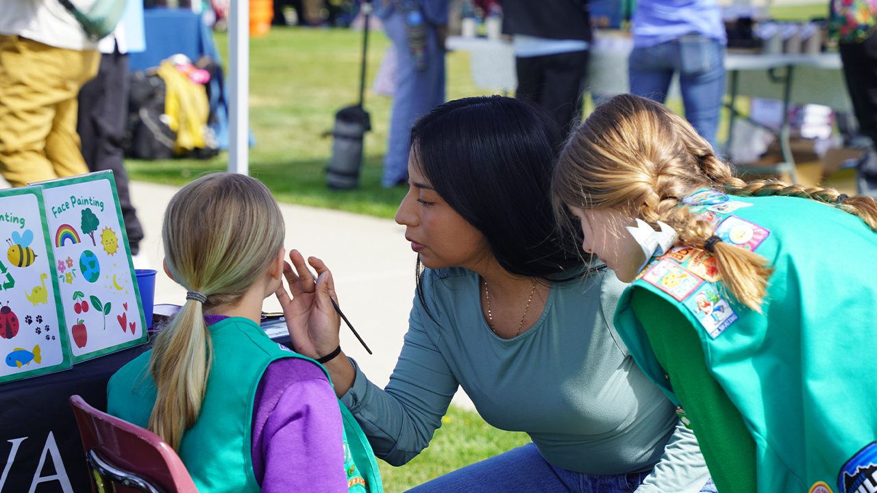 A WVC staff member kneels down and paints a child's face at the Sustainable NCW Earth Day Fair.