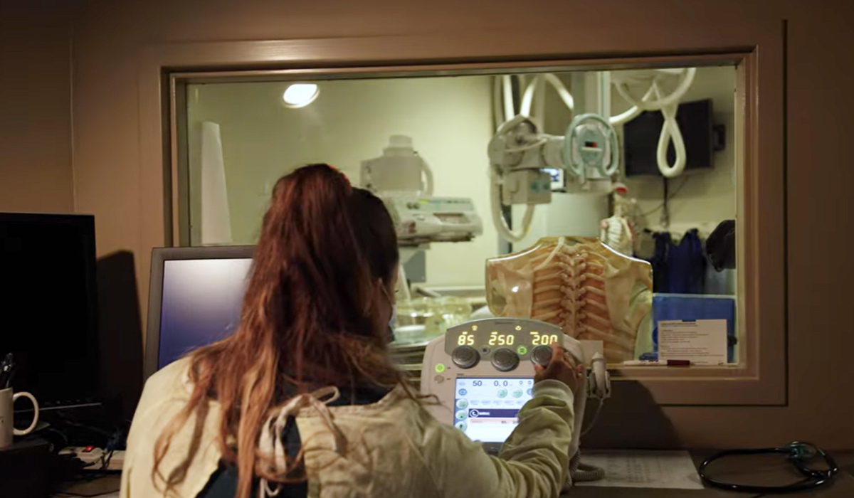 A radiologic technology student sits at a computer in a dark room while an instrument scans a bone.