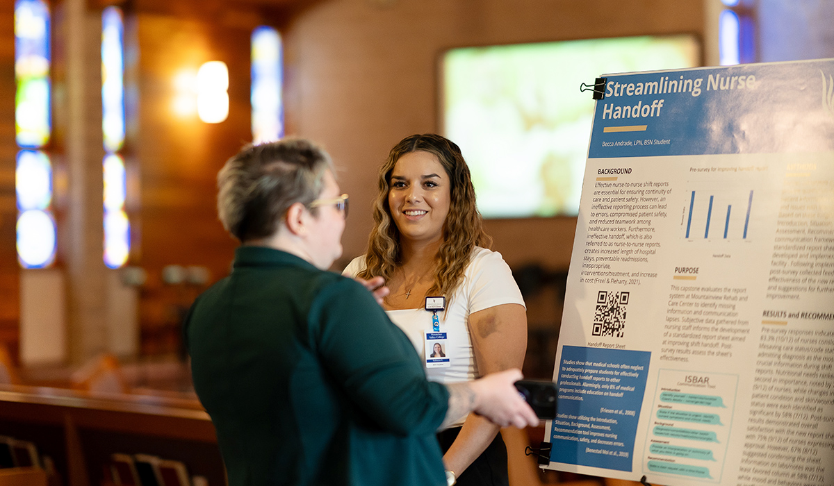 An LPN to BSN graduate stands next to her research poster while a professor observes it.