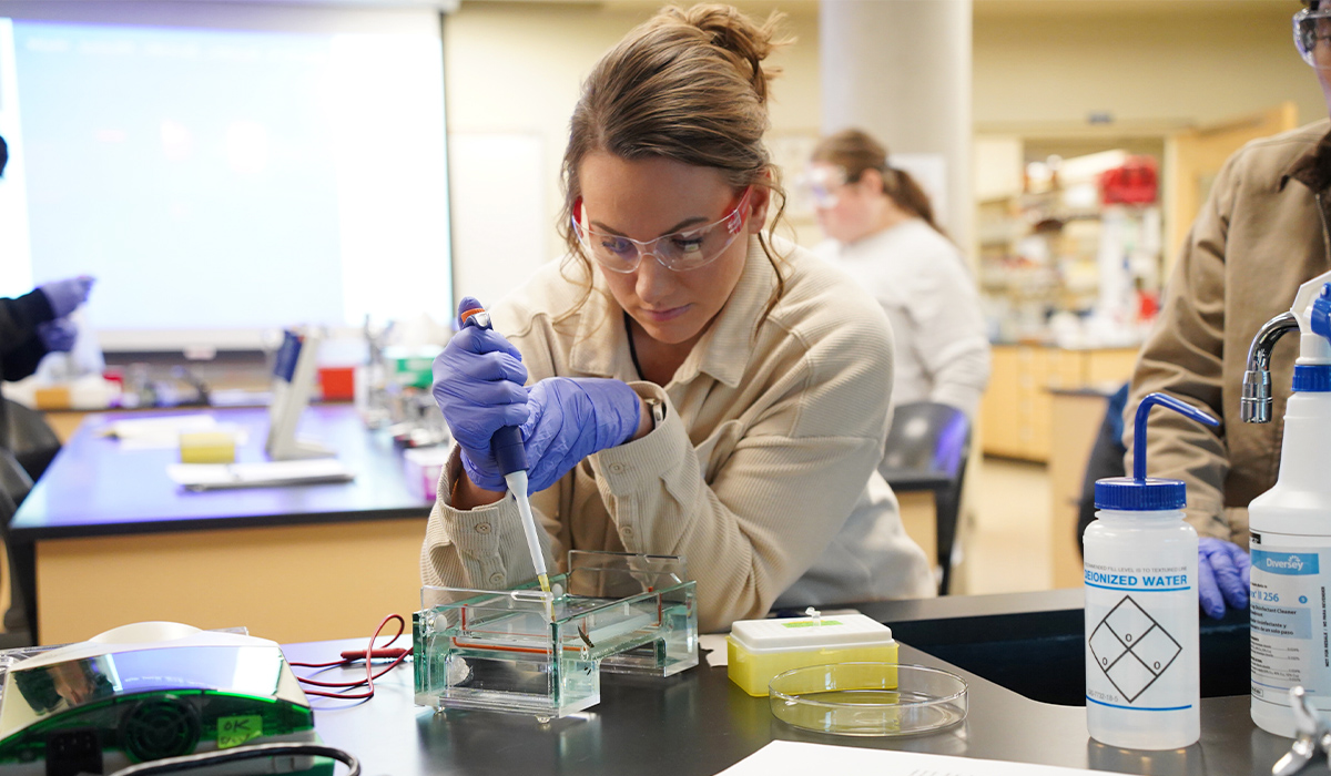 A biology student wearing a lab coat and protective glasses conducts an experiment in a lab.