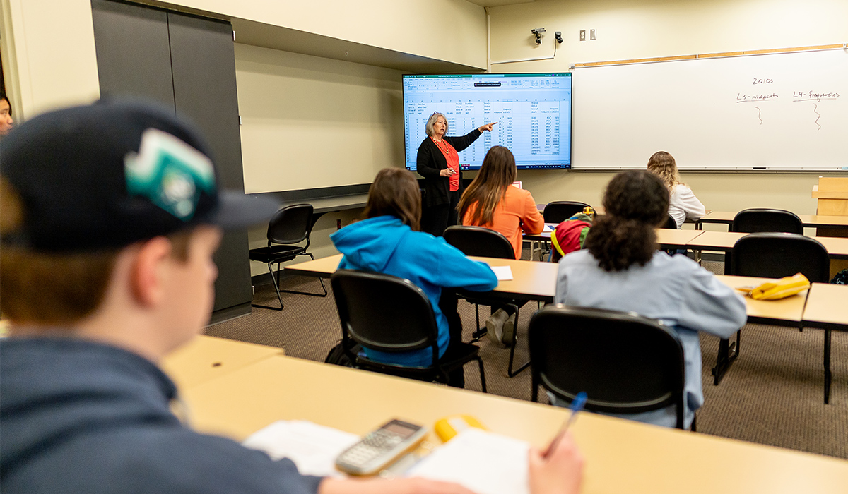 A math instructor stands at the front of a classroom with students looking on.