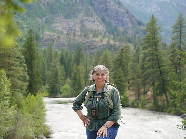 Heather Wallis Murphy stands in the foreground with a river, forest, and mountains behind her.