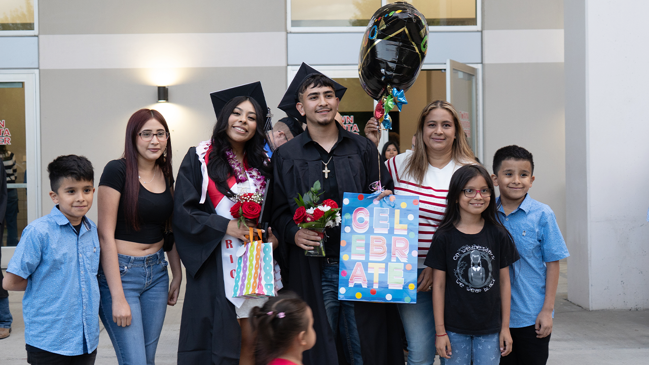 Two graduates stand with their family members and hold flowers and balloons after the graduation ceremony.