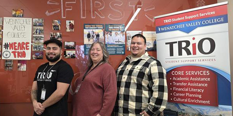 Three TRIO staff members stand together with the TRIO banner next to them.