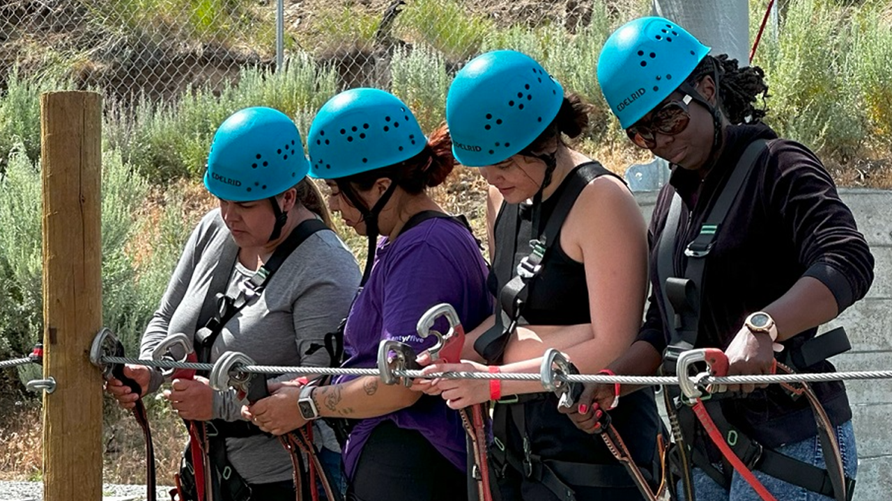 Four TRIO students wearing blue helmets and safety gear attach zipline clips to a wire.